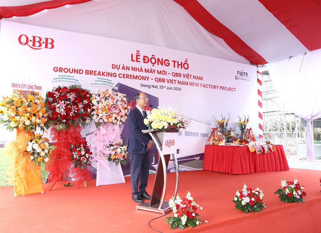 Người phát biểu đứng tại bục có hoa trang trí trước backdrop QBB Việt Nam.