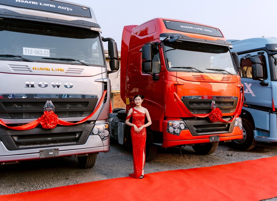 PG mặc đầm đỏ đứng tại khu vực trưng bày xe Hoàng Long Auto cạnh thảm đỏ.
